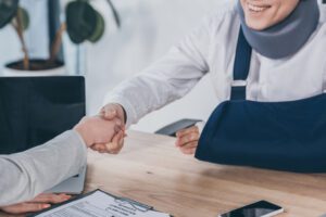 cropped view of woman shaking hands with worker in neck brace and arm bandage over table in office
