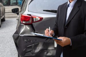 Side view of insurance officer writing on clipboard while insurance agent examining black car after accident.